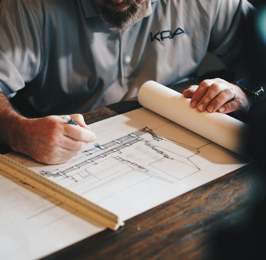 Person sketching architectural plans on paper at a wooden table, with rolled blueprints nearby