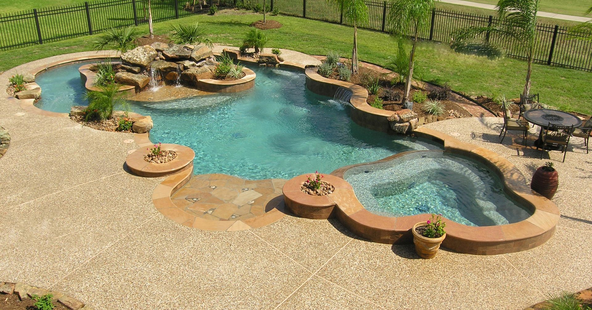 Backyard pool with curving tan stone edging and turquoise water, surrounded by gravel and green lawn.