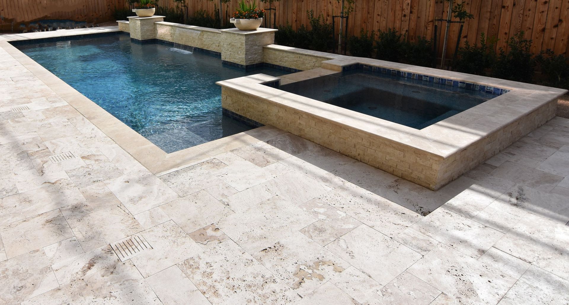 Backyard pool with attached square hot tub, surrounded by light stone patio and wooden fence