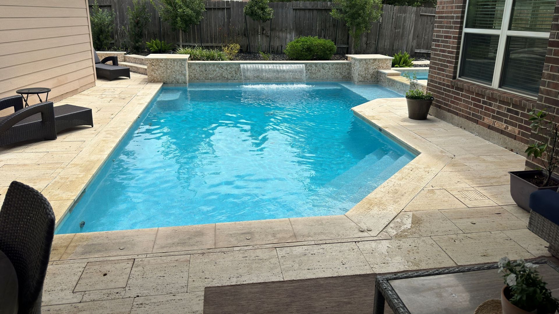 Backyard swimming pool with blue water, surrounded by a beige stone patio and brick house exterior.