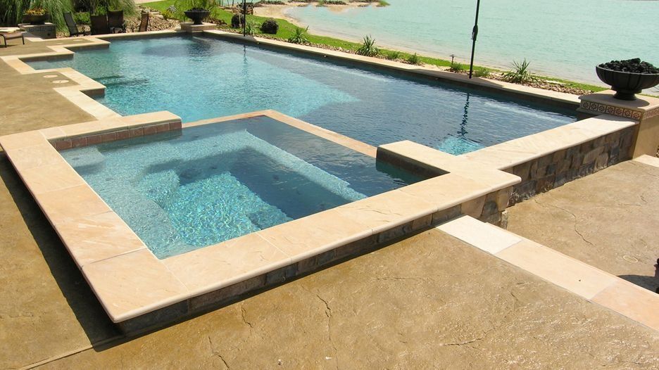 Outdoor pool with a raised square hot tub and tan stone deck overlooking a body of water