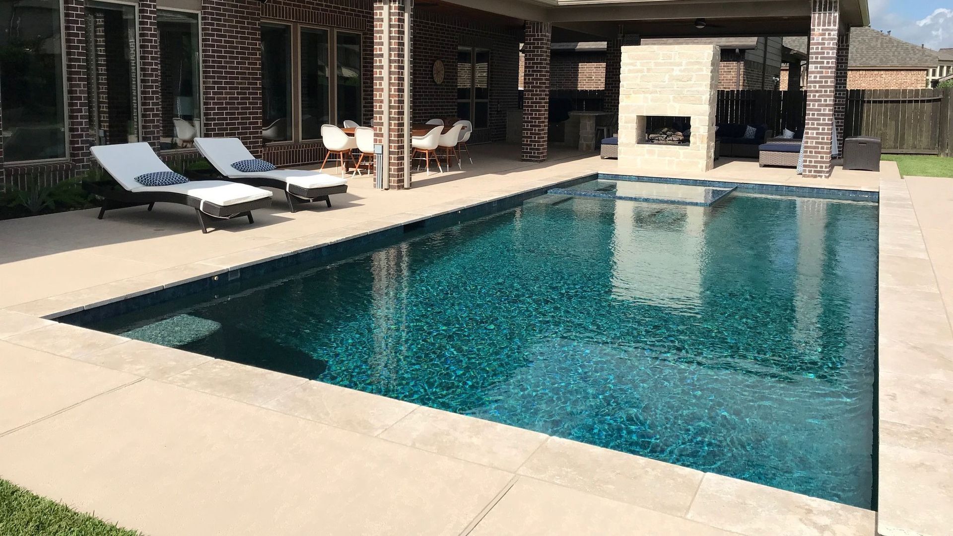 Modern backyard pool with blue water, lounge chairs, and a covered patio beside a stone fireplace