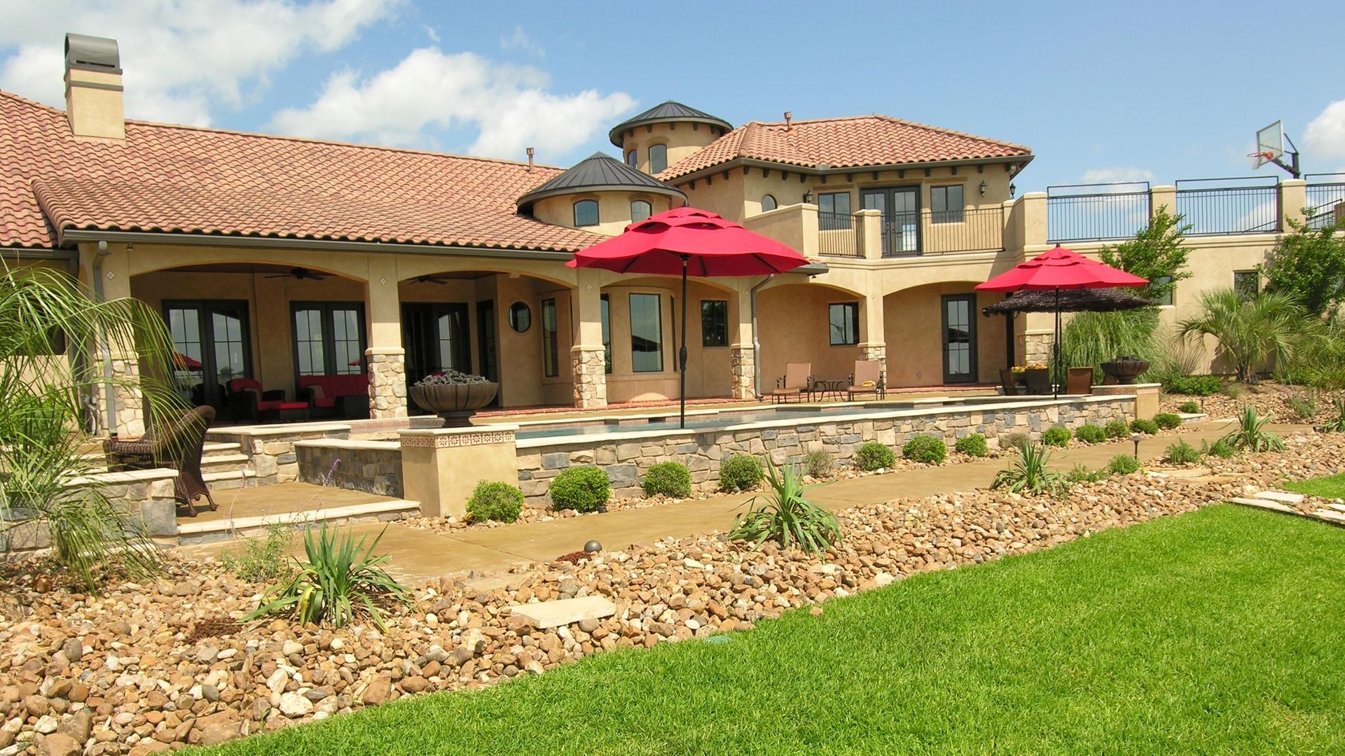 Sunlit Mediterranean-style house with red umbrellas, stone patio, and green lawn in a landscaped backyard