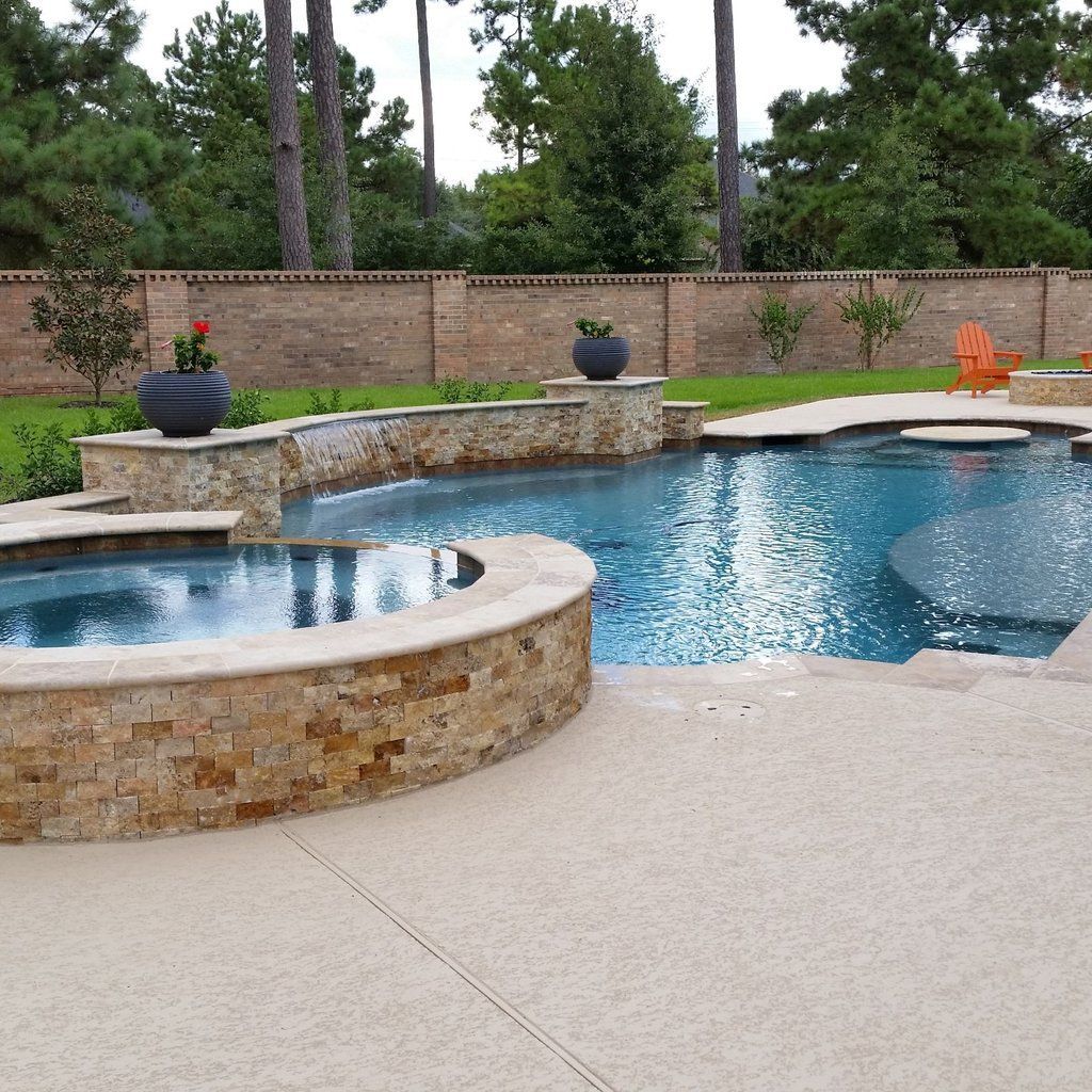 Backyard pool with stone spa, curved blue water, and a tan deck bordered by a brick wall and trees