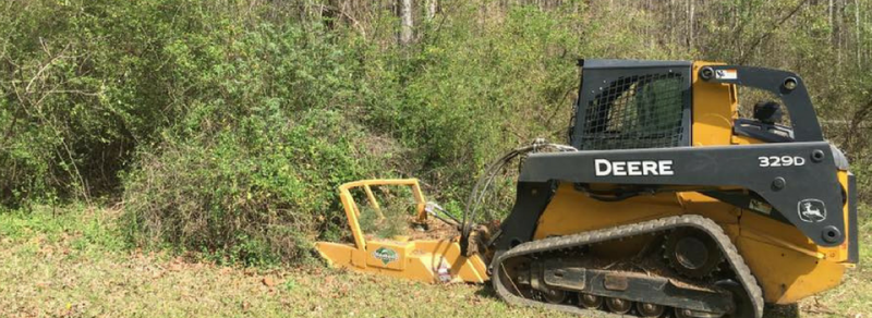 A yellow John Deere compact track loader with a brush cutter attachment clears thick green vegetation in a wooded area.