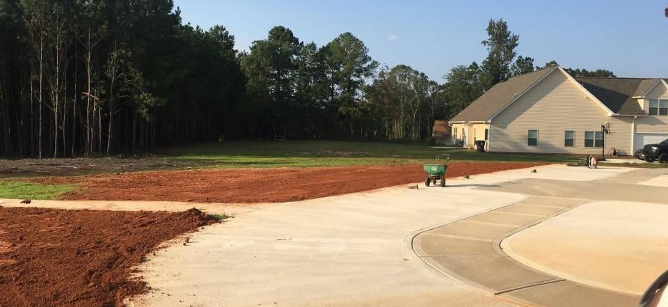 A construction site with red soil, a concrete driveway, a green wheelbarrow, and a house bordering a forest.