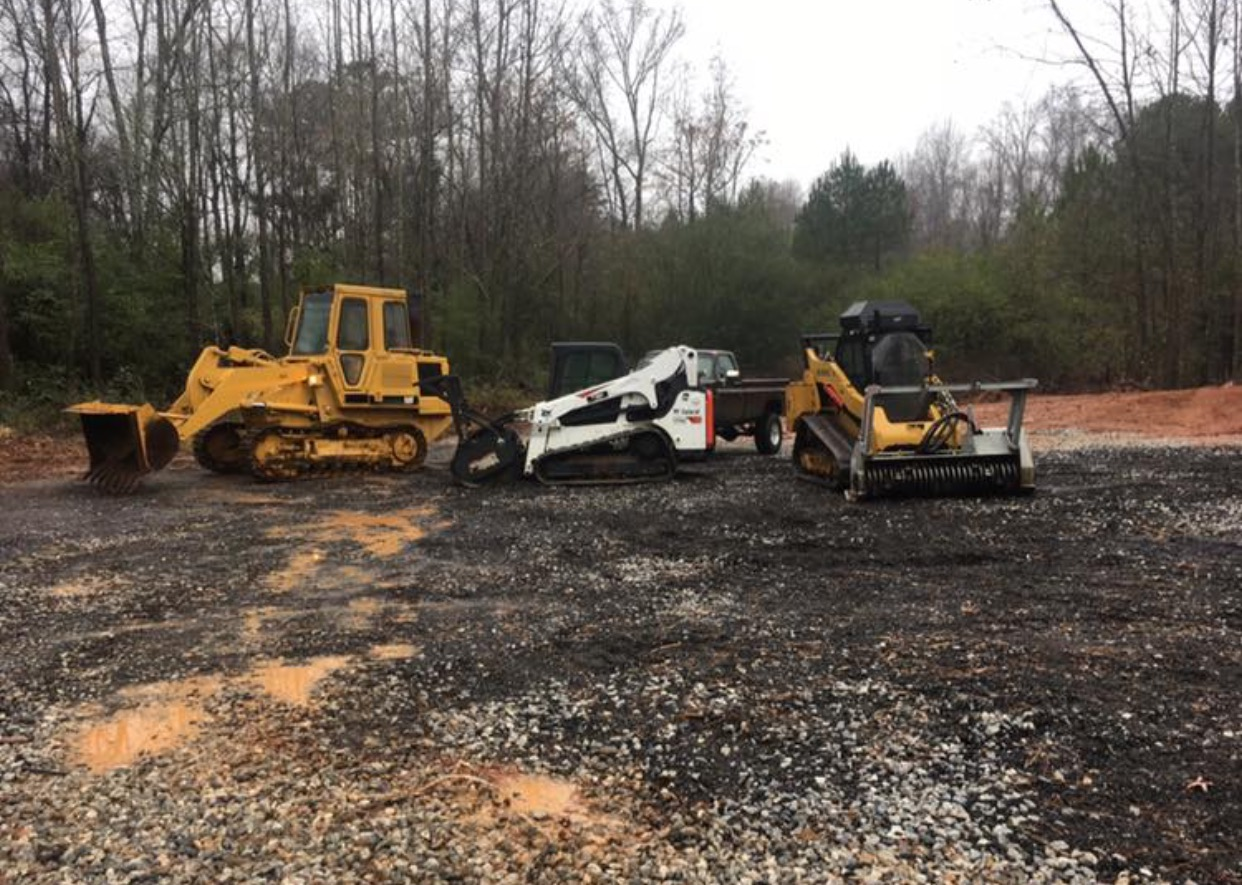 Three construction vehicles parked on a gravel lot in front of a tree line on a cloudy day.