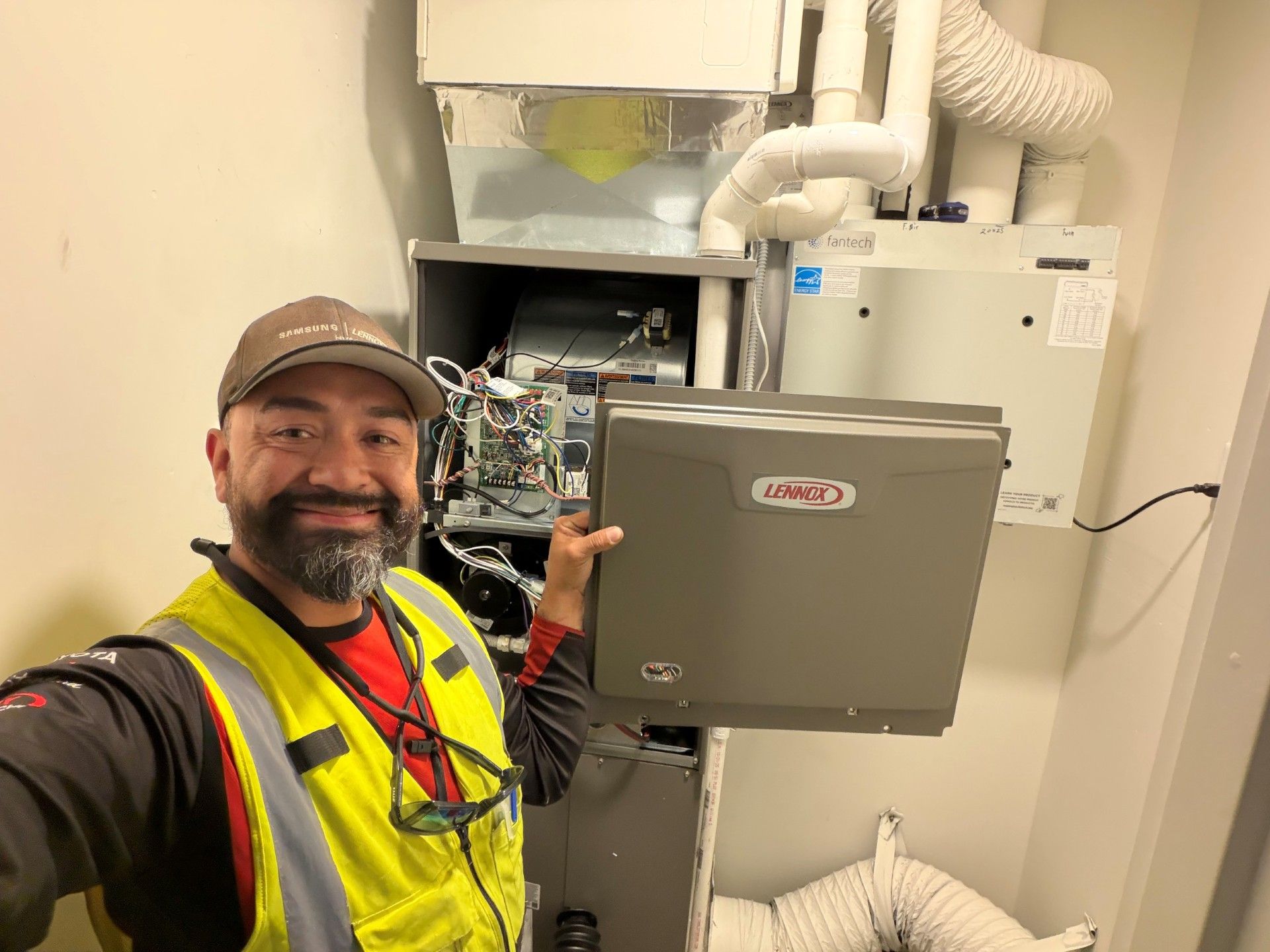 Technician smiling beside open HVAC equipment in a utility room, wearing a cap and safety vest