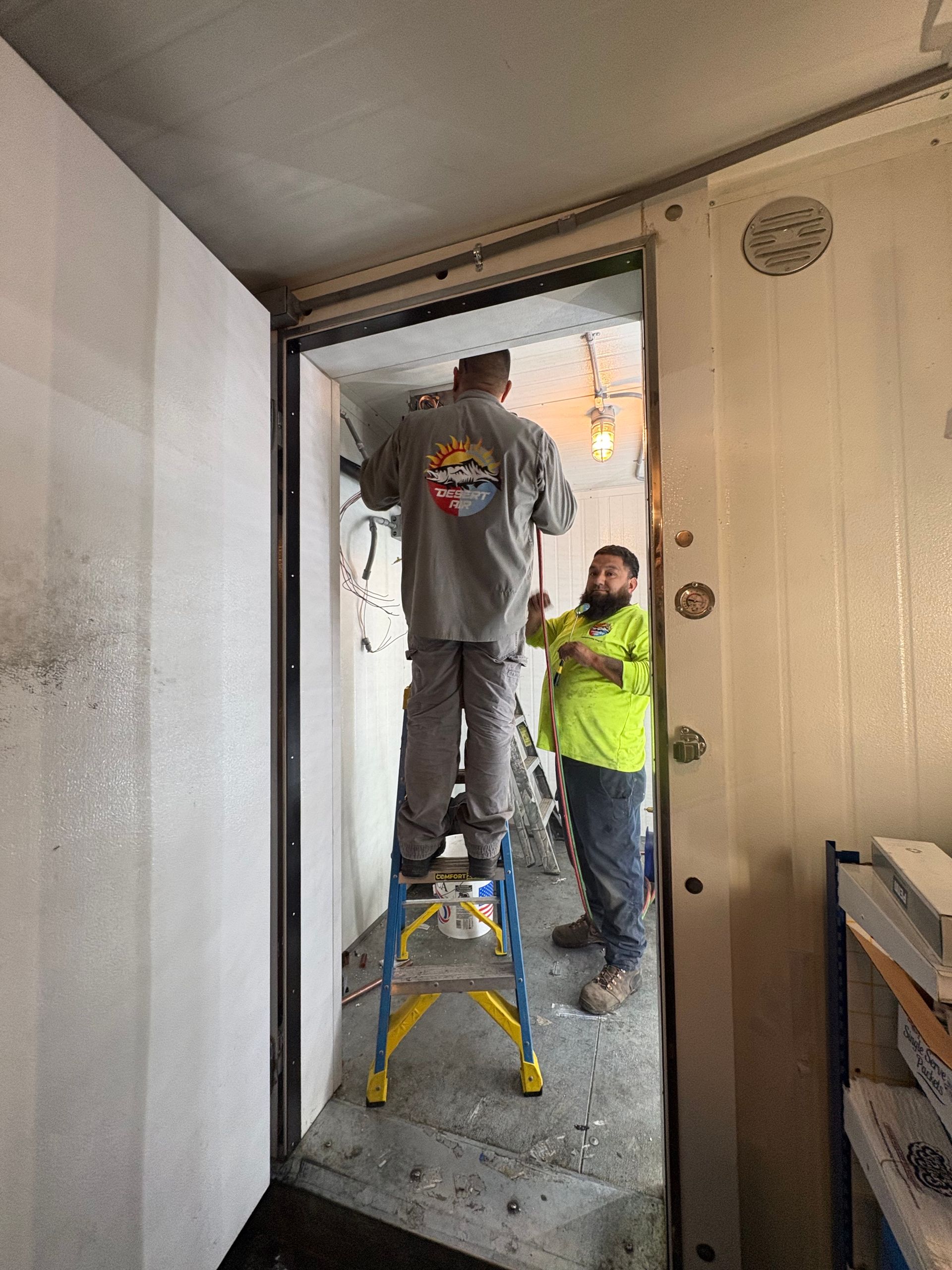 Two workers repairing a doorway, one on a step stool and one holding a tool inside a hallway.