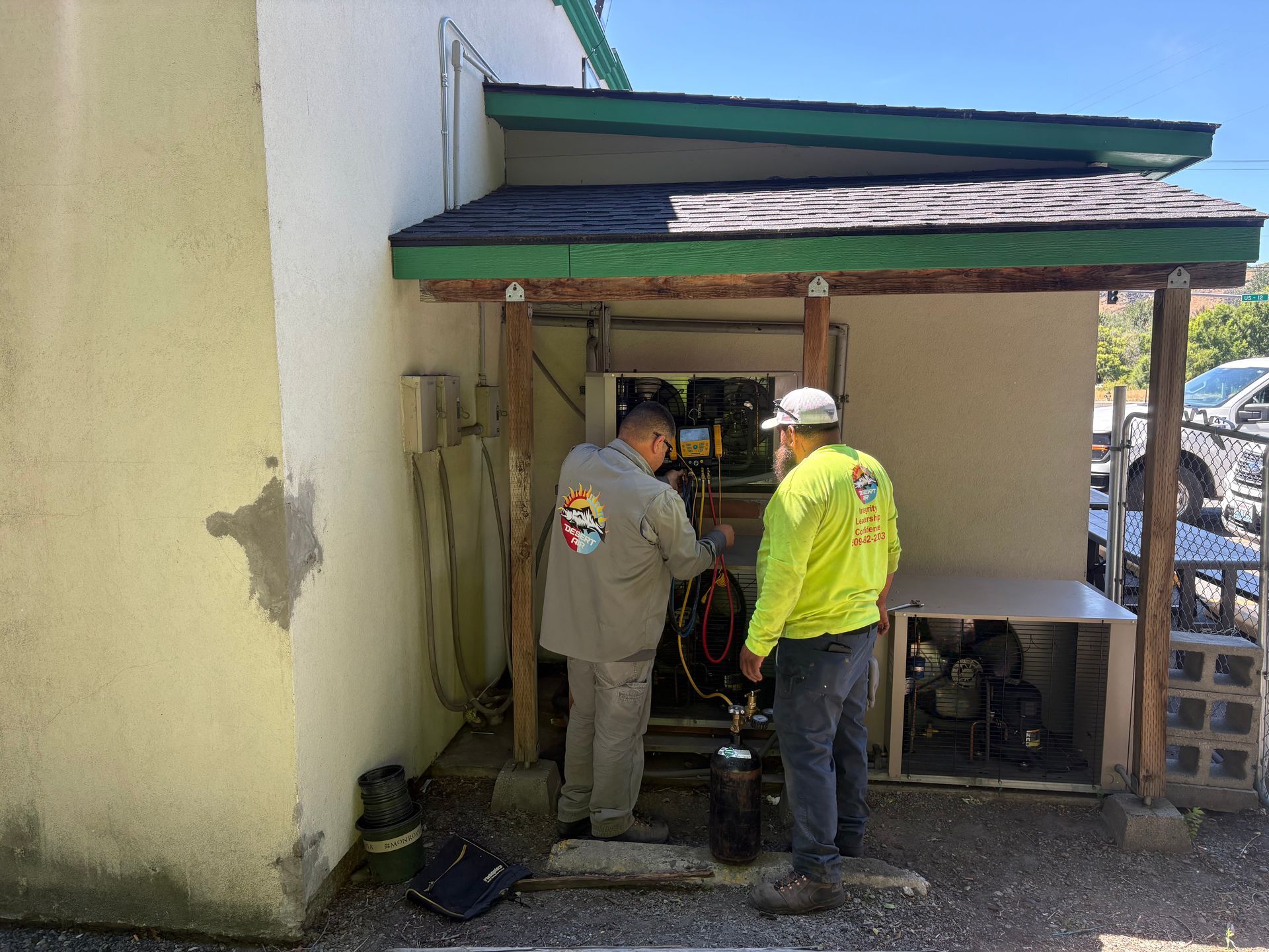 Two workers inspecting equipment beside a small building under a green awning.