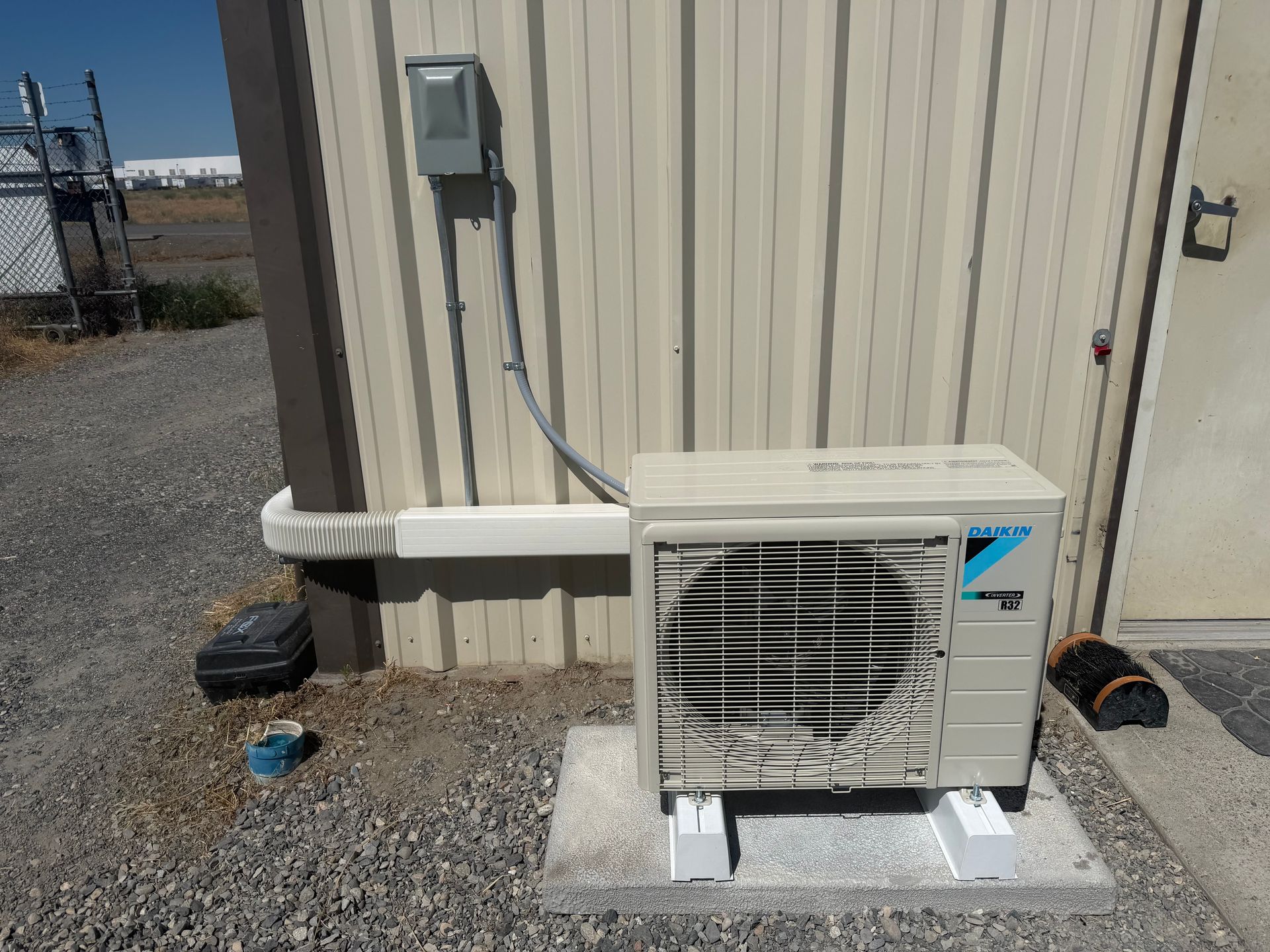 Outdoor HVAC unit beside a beige metal building with electrical conduit and gravel ground