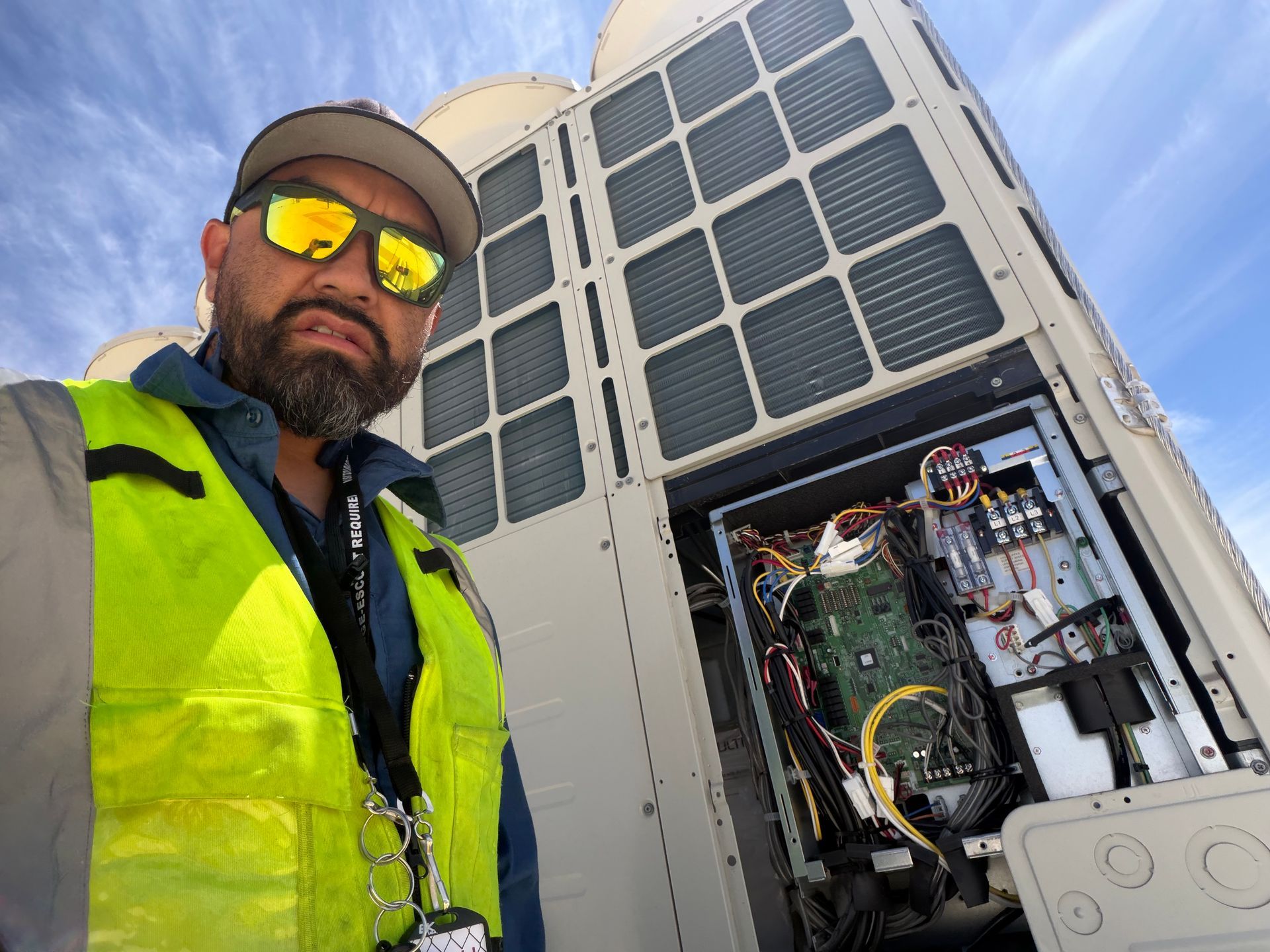 Technician in a neon vest standing beside an open HVAC unit with exposed wiring outdoors.