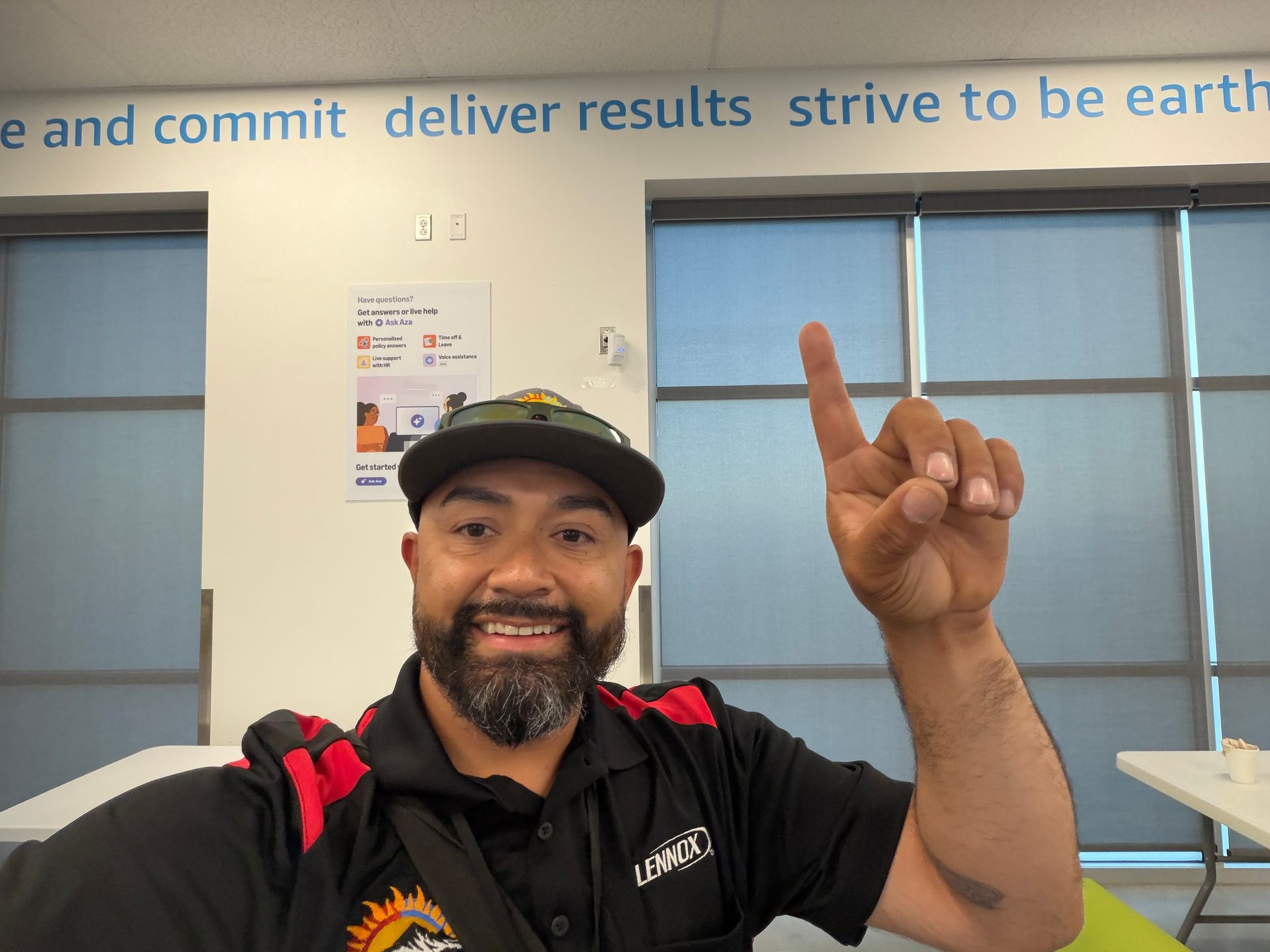 Man smiling in a black shirt and cap, pointing upward in an office with windows and wall text.