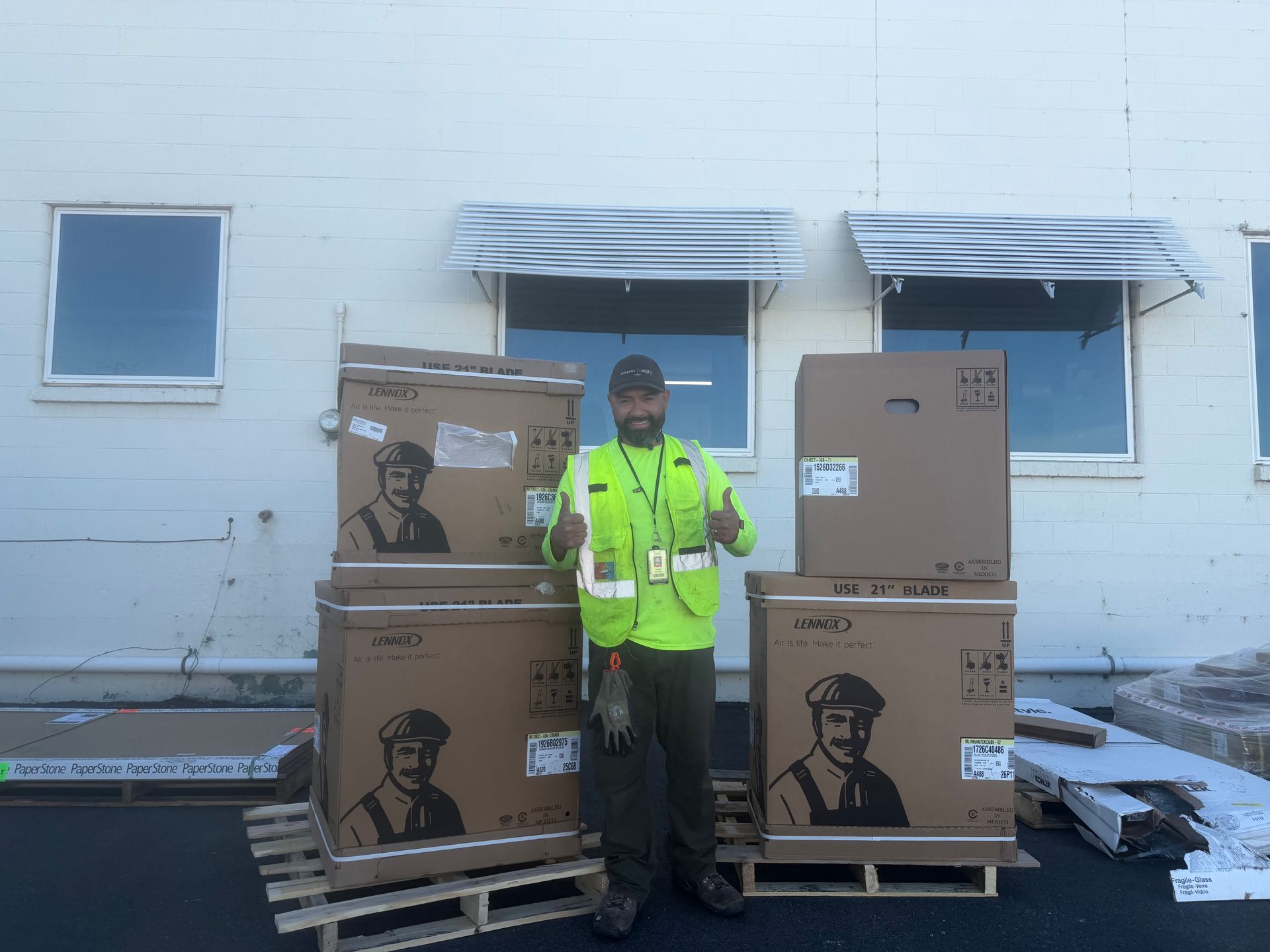 Worker in neon safety jacket standing between stacked cardboard boxes outside a building with damaged windows.