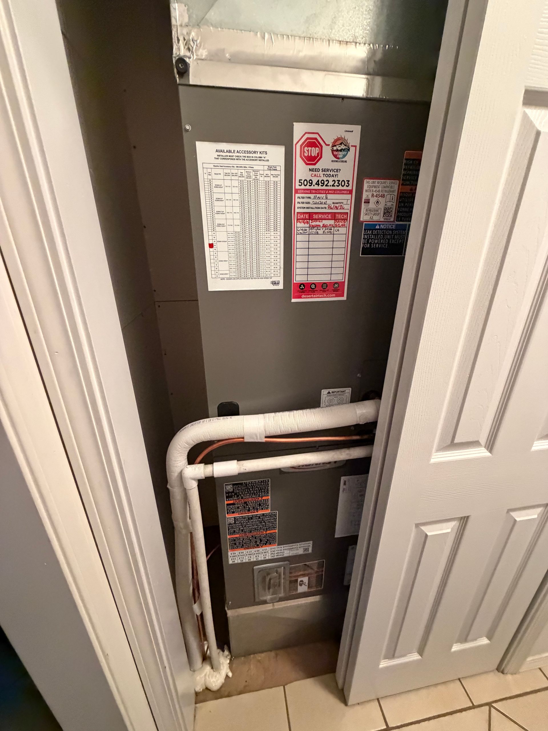 Open HVAC closet with gray furnace, white pipes, and inspection labels behind a partly open door