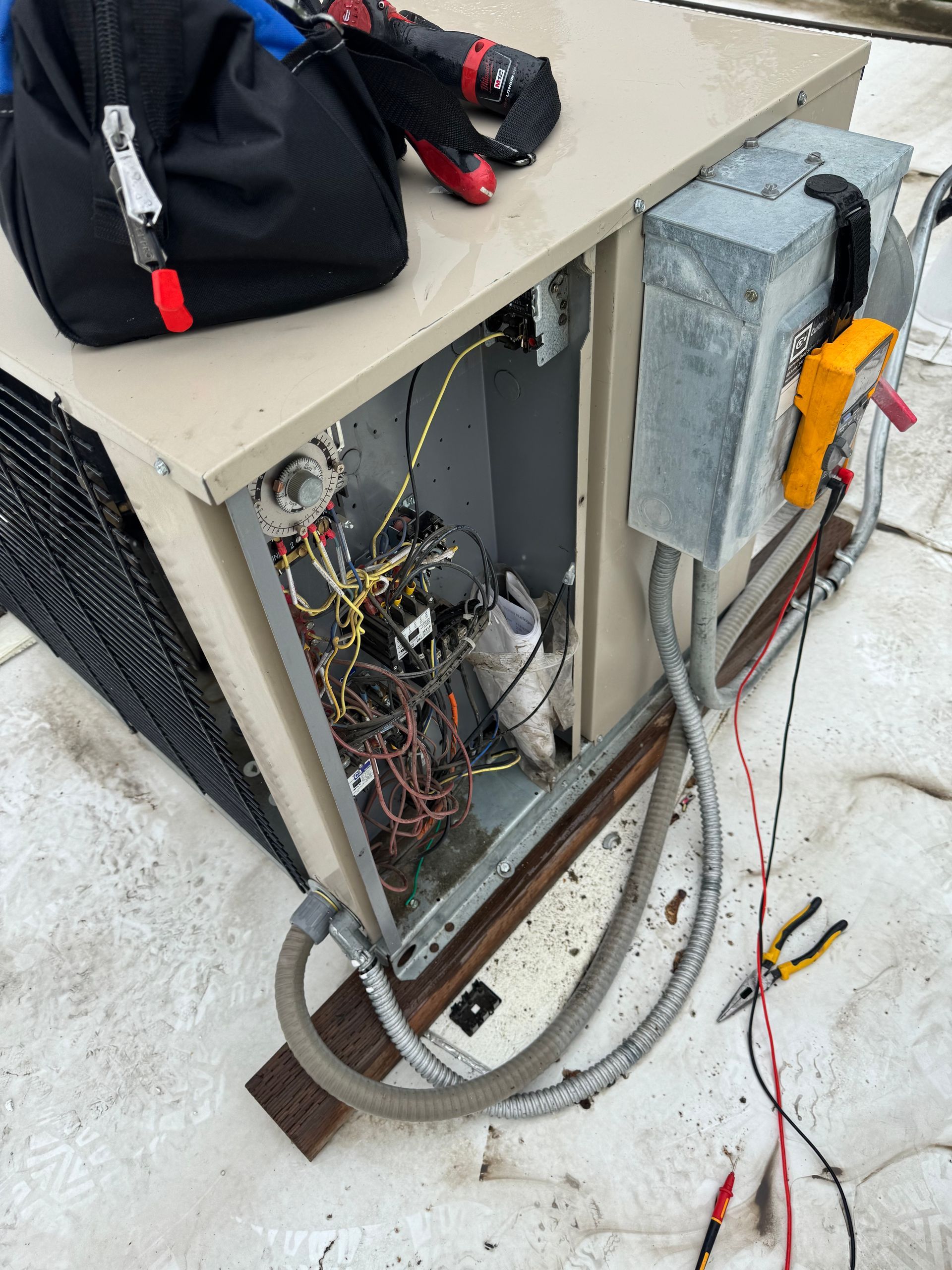 Open electrical cabinet with exposed wiring, meter, and cables beside tools on a snowy ground