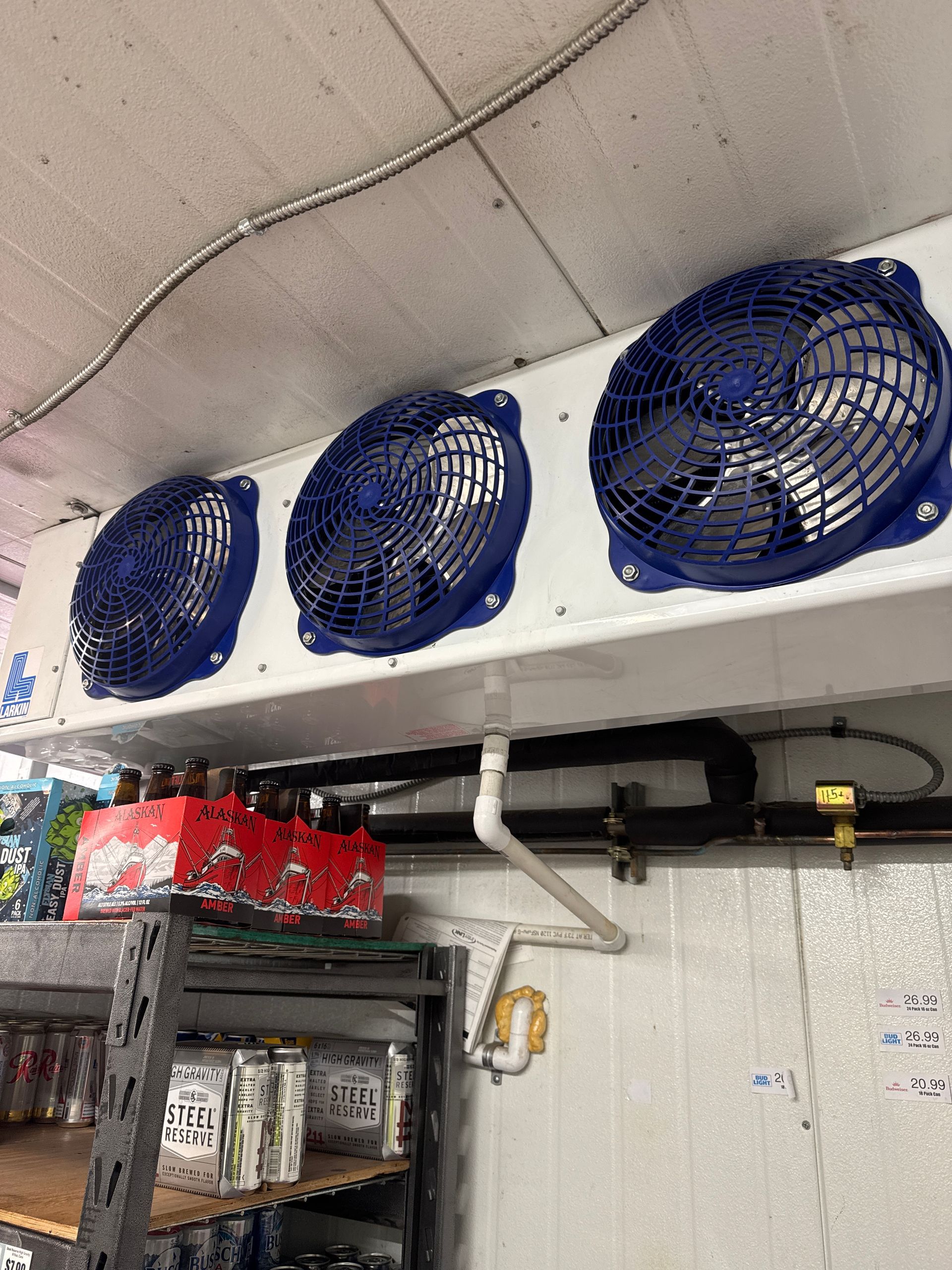 Wall-mounted white cooler with three blue fans above store shelves and beverages