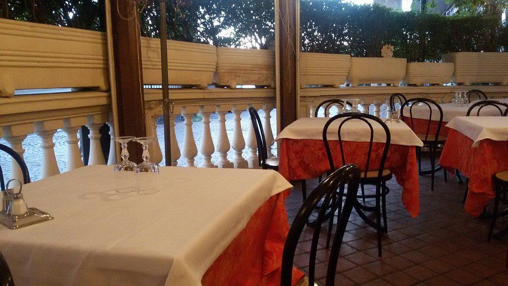 Tavolini vuoti di un caffè all'aperto, con tovaglie bianche e gonne rosse, accanto a un patio con vetrate al crepuscolo.