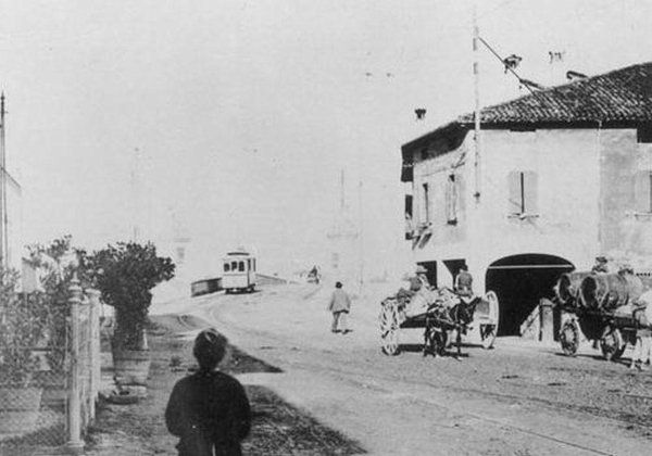 Scena di strada d'epoca con carrozza trainata da cavalli, pedoni e un tram vicino a un edificio d'angolo.