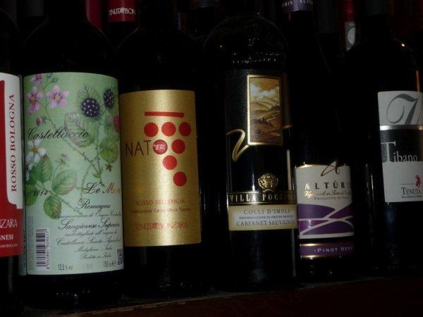 Diverse bottiglie di vino allineate su uno scaffale, con etichette scure e una con etichetta dorata decorata con cerchi rossi.