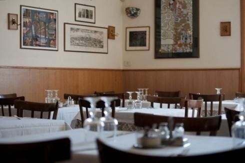 Sala da pranzo vuota di un ristorante con tovaglie bianche, sedie scure e quadri incorniciati su pareti rivestite in legno.