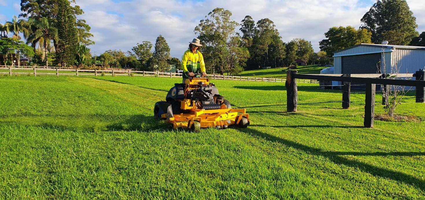 A Man Is Riding a Yellow Lawn Mower on A Lush Green Field — All Round Ground Maintenance in Woolgoolga, NSW