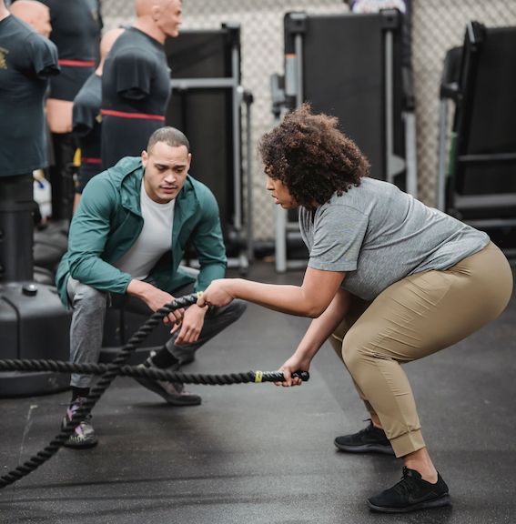 Two people doing battle rope exercises in a gym, one crouching in teal and one pulling in gray and tan.