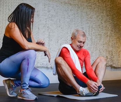 Fitness instructor kneels beside a man stretching on a mat in a studio, offering guidance.