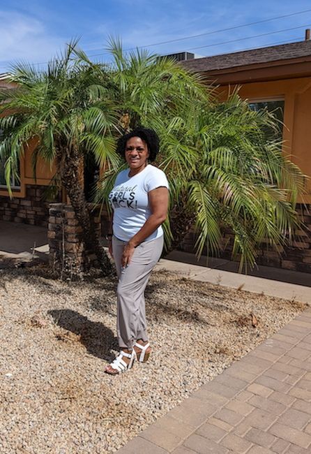Shvonne standing by a palm tree in a sunny courtyard, wearing a white T-shirt and gray pants.