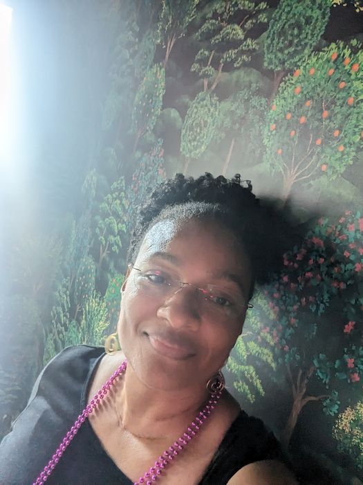 Shvonne smiling with glasses and purple beads, standing by a floral wall mural