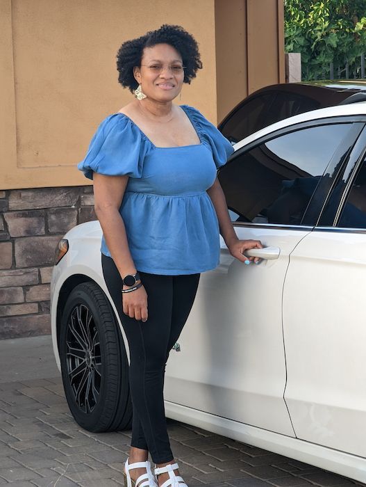 Shvonne standing beside a white car, wearing a blue blouse and black pants, smiling outdoors.