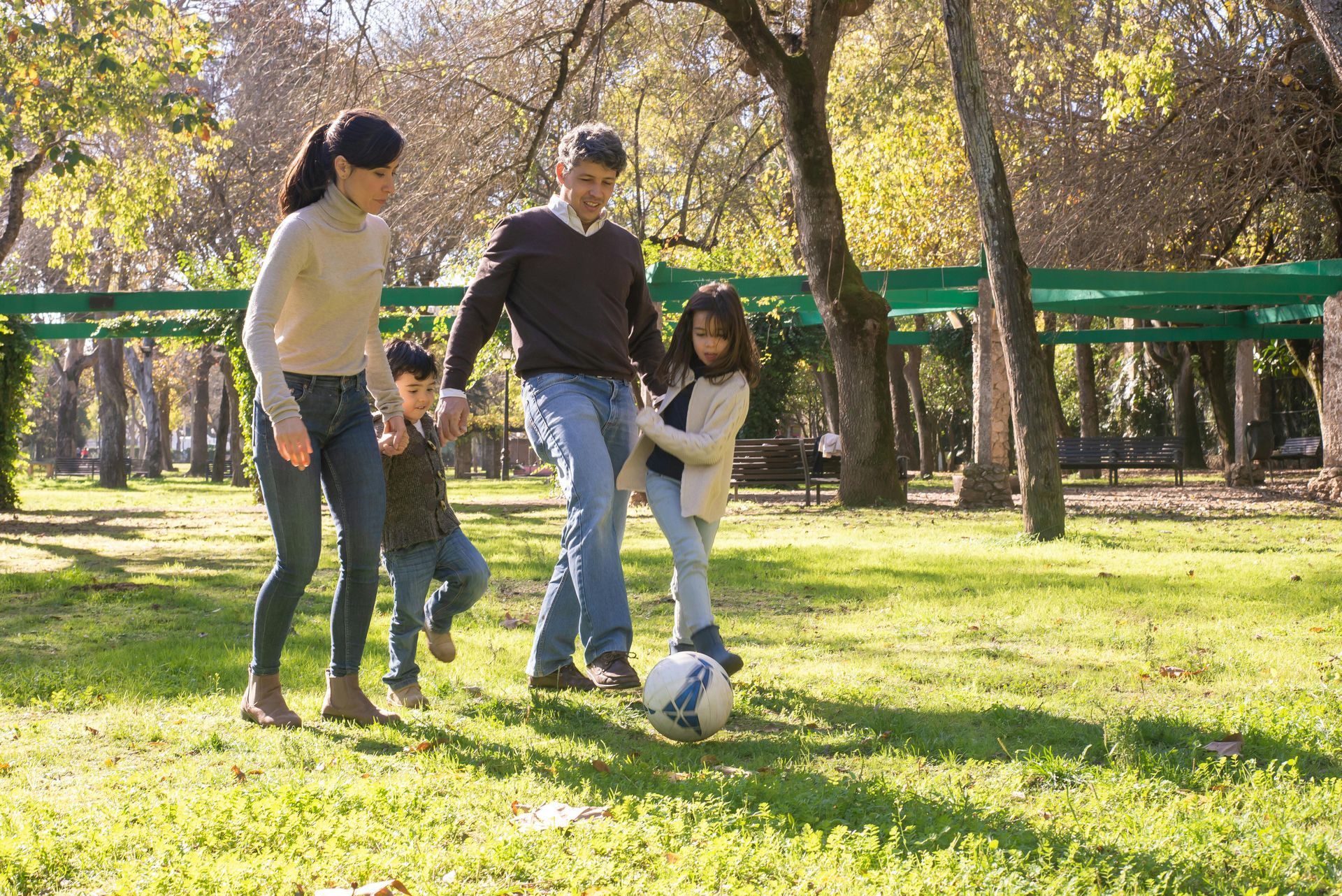 Four people playing soccer in a sunny park, with a ball on the grass and trees in the background