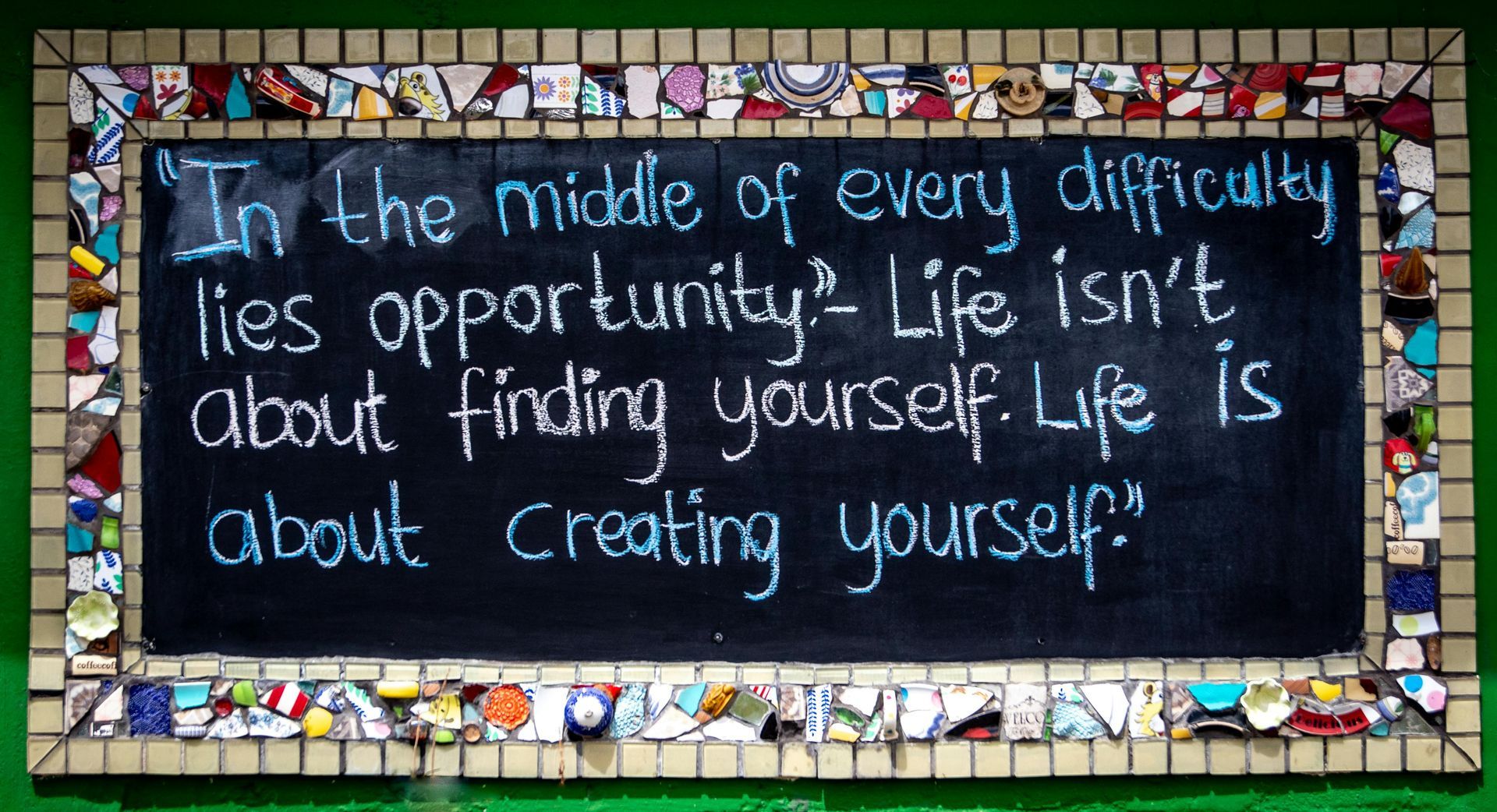 Chalkboard sign with blue handwritten quote framed by colorful mosaic tiles.