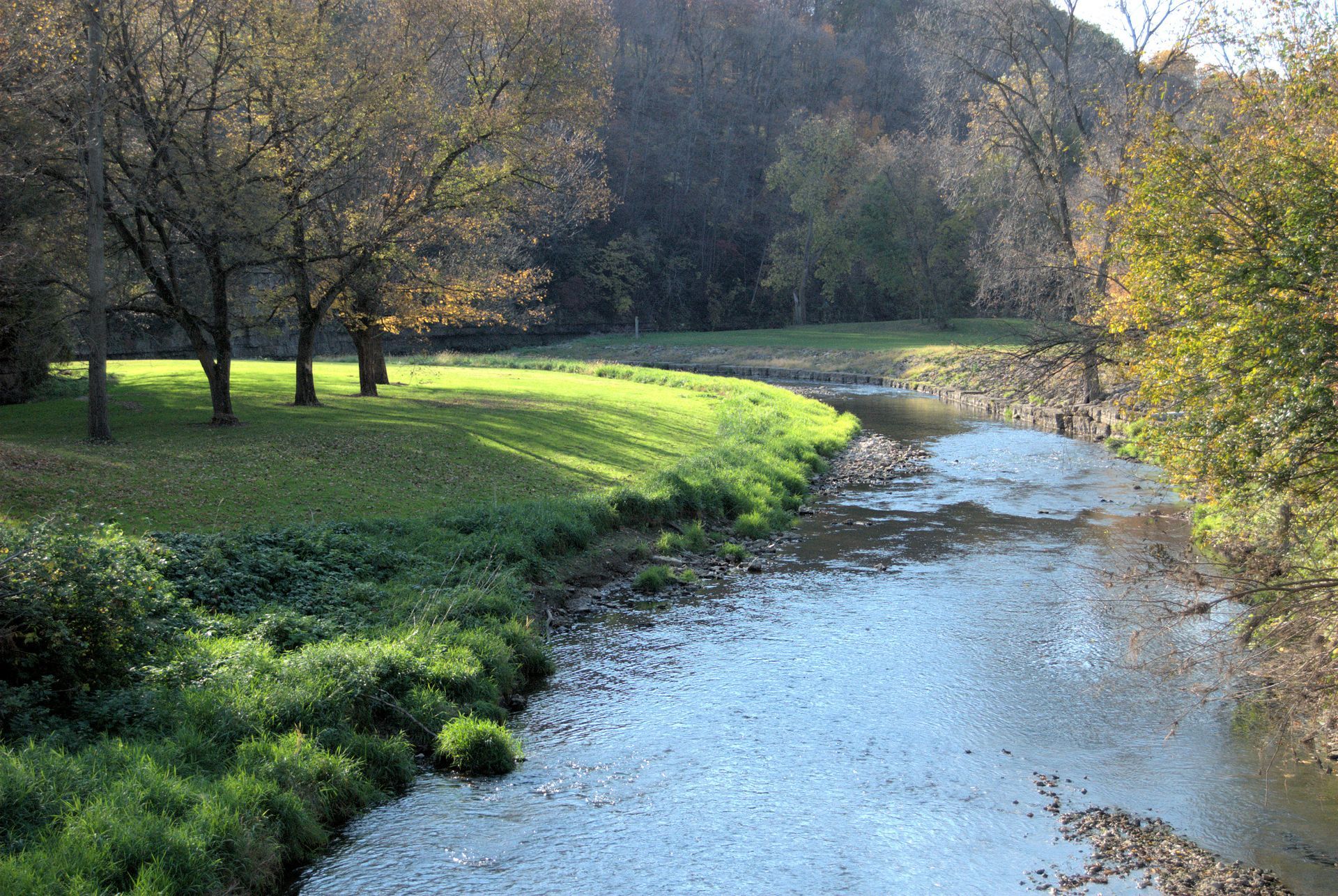 Sunlit river curving through a grassy park with trees and hills in the background