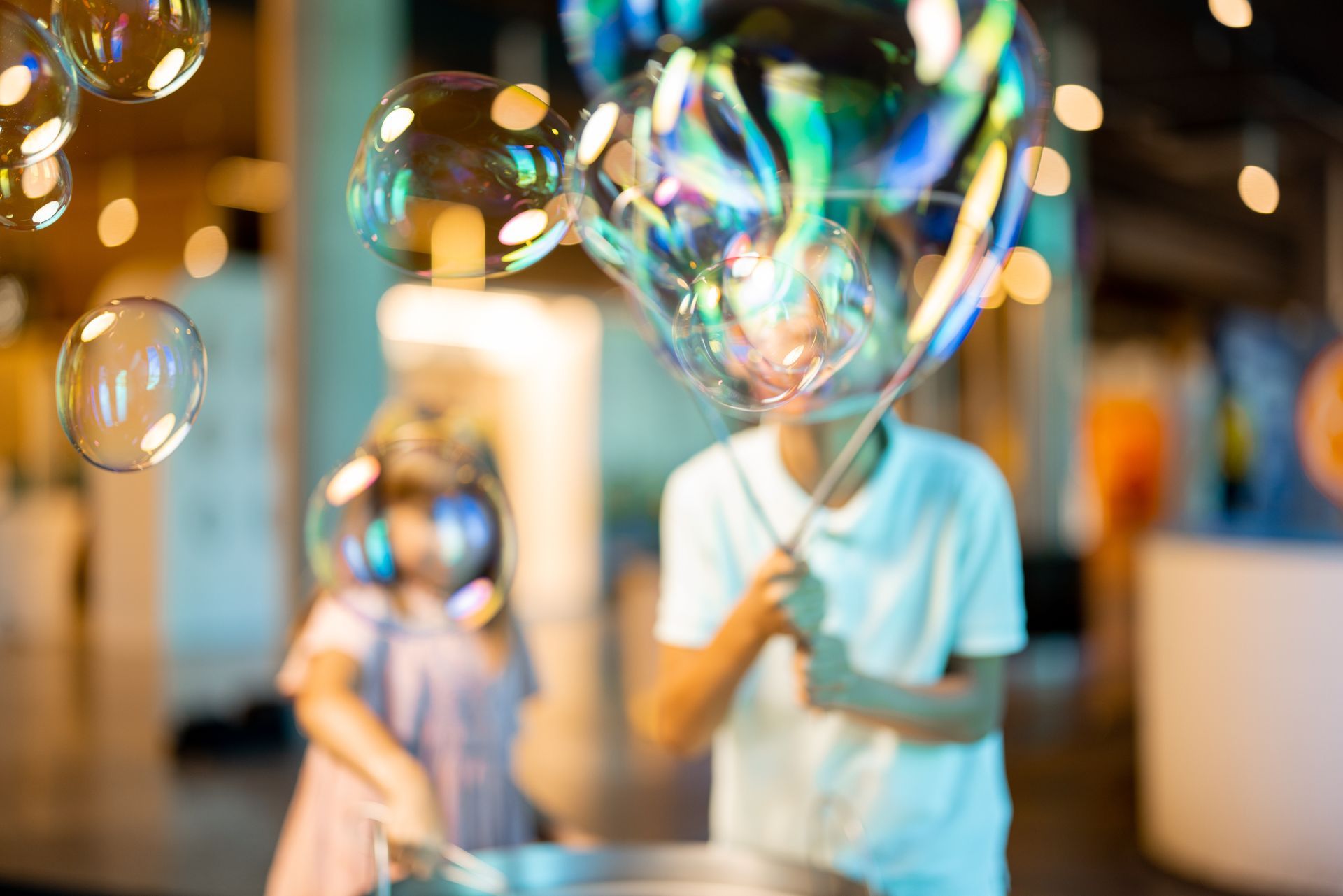 Two children blowing large, iridescent bubbles indoors.