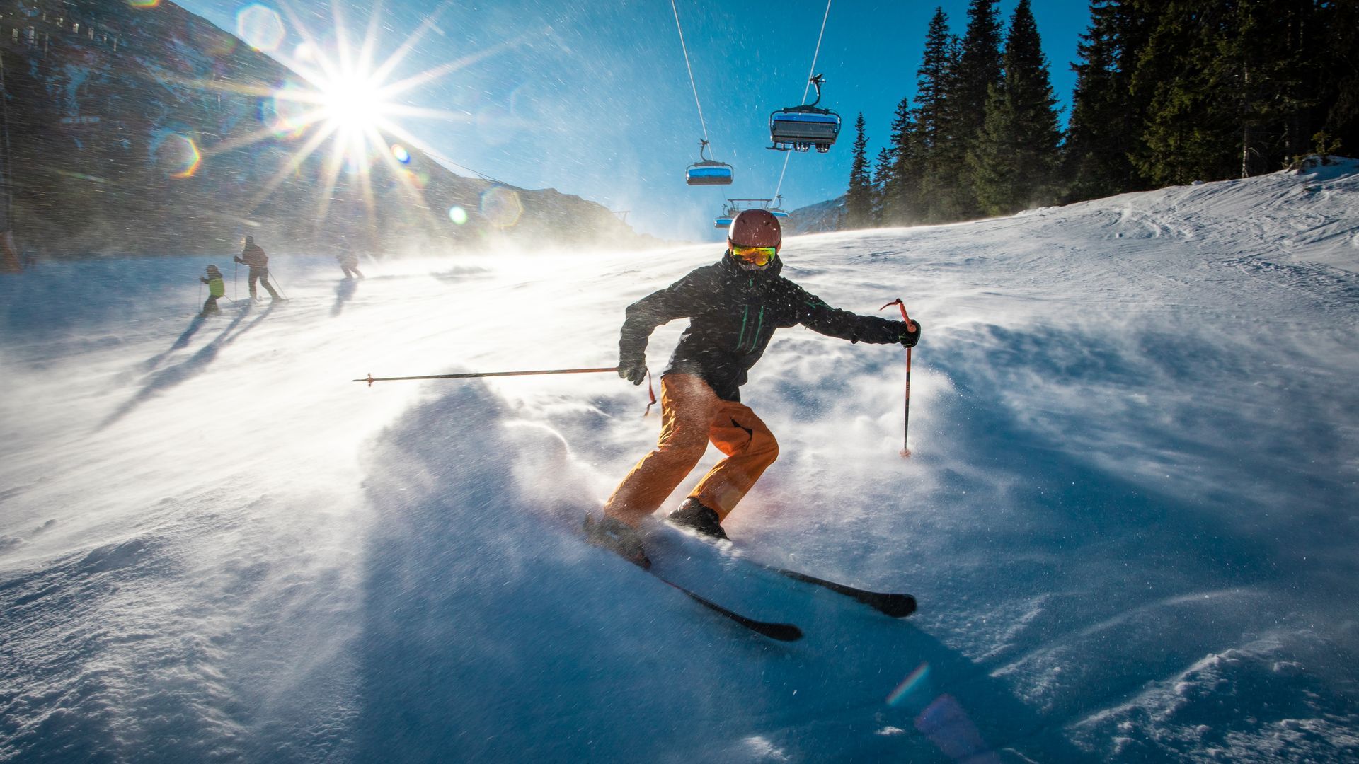 Skier carving through fresh snow under bright sun, with chairlift and pine trees in the background