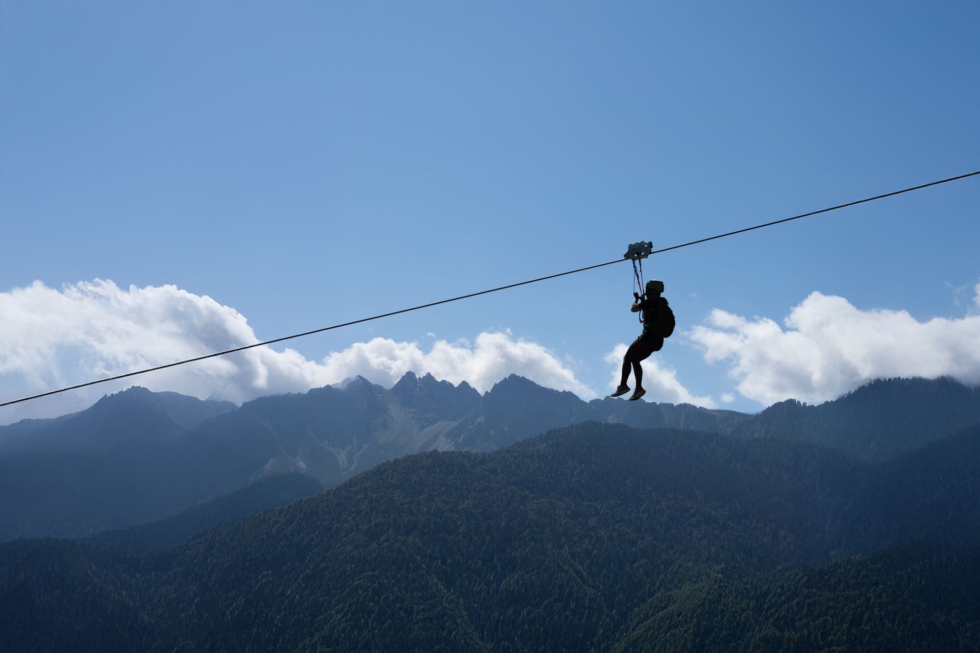 Person zip-lining above a forested mountain valley under a blue sky