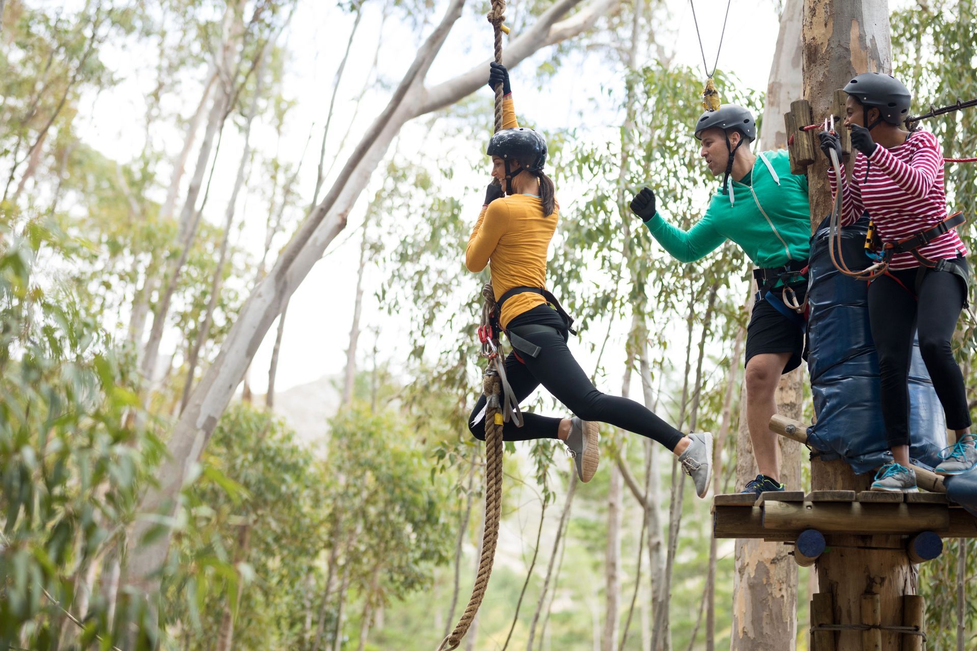 Three people in helmets on a ropes course platform, one stepping off into a zipline in a wooded area.