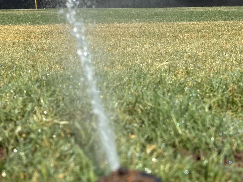 A sprinkler sprays water over a green grassy field in sunlight.