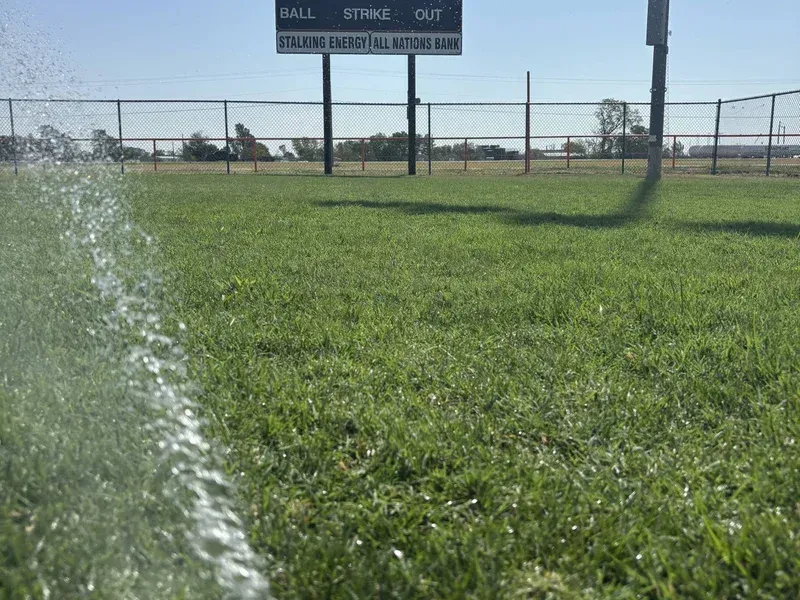 Sprinkler spraying grass on a sunny sports field with a scoreboard and fence in the background