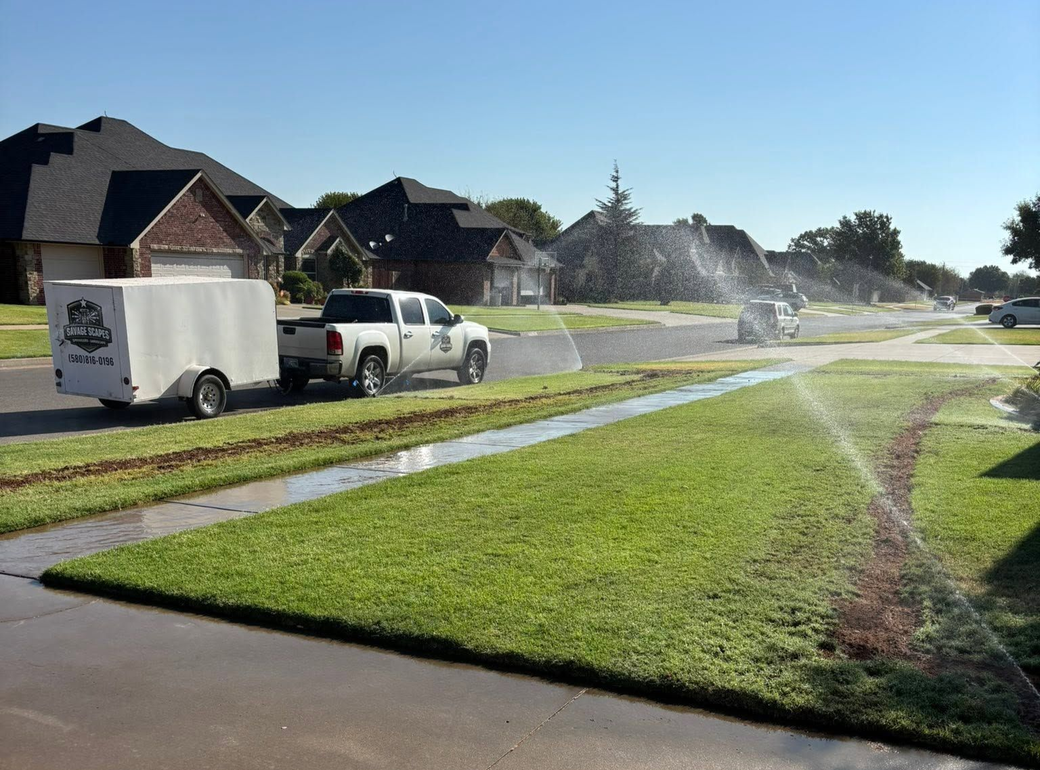 Sprinklers watering a suburban street lawn beside parked pickup trucks and houses on a sunny day