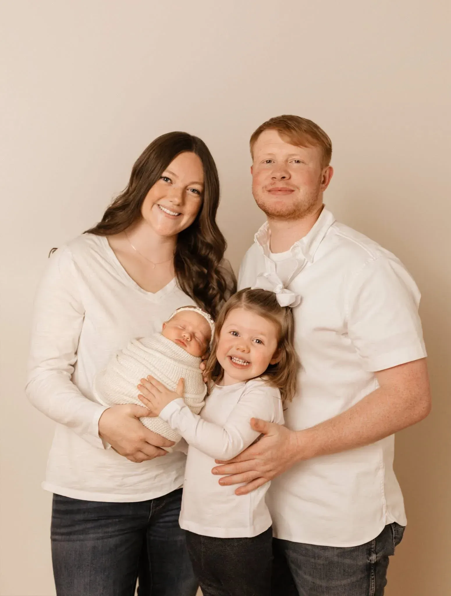 Family of four posing together against a plain beige background, smiling and holding a baby.