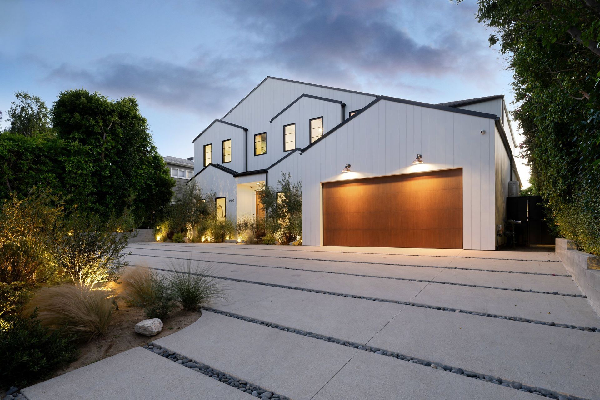 Modern white two-story house with lit garage and sloped driveway at dusk
