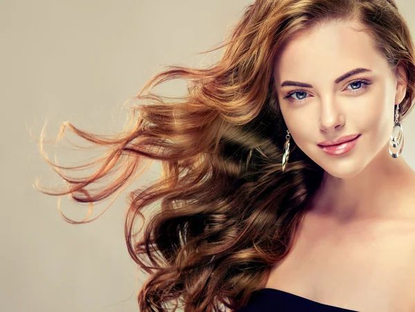 Una donna con i capelli castani ondulati sorride, i capelli sciolti. Indossa orecchini, un top nero e uno sfondo neutro.