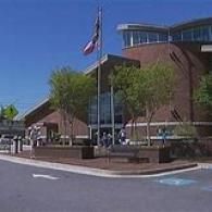 A modern red-brick building with curved glass architecture, a flag flying, and a parking lot entrance on a sunny day.