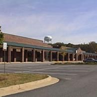 A long, single-story brick commercial building with a covered walkway and parking lot, with a water tower in the distance.