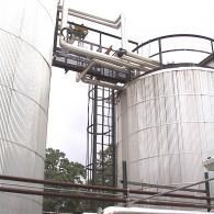 Two large, silver industrial storage tanks connected by an elevated metal walkway and ladder against a bright sky.