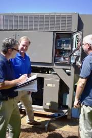 Three people stand outdoors, gathered around an open industrial HVAC unit, reviewing technical documents.