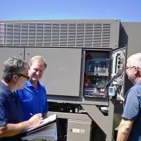 Three people standing outdoors examining the open electrical control panel of a large industrial HVAC unit.