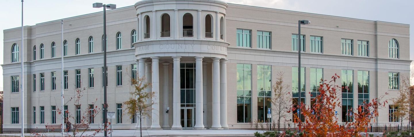 A multi-story, beige stone building featuring a central portico with columns and arched windows, surrounded by trees.